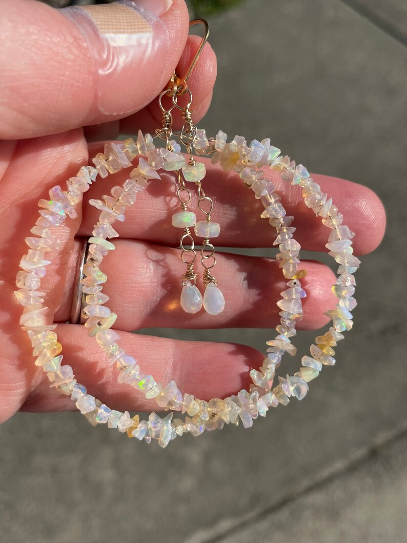 May include: A pair of gold hoop earrings with a double row of iridescent white opal chips. The earrings have a dangling drop of opal at the bottom.