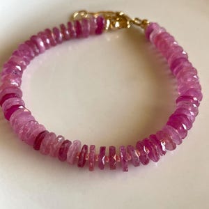 May include: A pink gemstone bracelet with a gold clasp. The bracelet is made of small, faceted pink gemstones. The gemstones are strung together to form a circular shape. The clasp is gold-coloured and secures the bracelet.