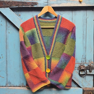Peut inclure: Un cardigan en tricot à col en V présentant un motif patchwork de carrés aux dégradés arc-en-ciel vibrants de rouge, orange, jaune, vert, bleu et violet. Le vêtement possède des manches longues, une bordure côtelée et trois gros boutons ronds unis vert lime et jaune.
