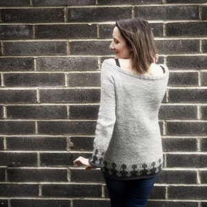 Puede incluir: Una mujer lleva un suéter gris y negro con un patrón geométrico en la parte inferior. Está de pie frente a una pared de ladrillo.