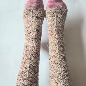May include: A pair of mid-calf length knit socks featuring a delicate, repeating scalloped lace pattern. The socks are crafted from a speckled beige yarn with dark brown flecks and have solid pink knitted toe sections and ribbed cuffs.