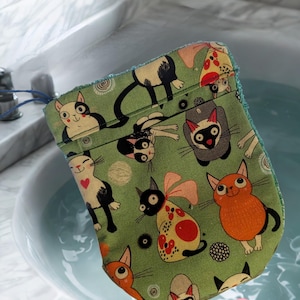 May include: A green cat-patterned washcloth with a blue drawstring, featuring cartoon cat illustrations in black, white, orange, and grey. The washcloth is in a bathtub with a pink bar of soap with the text "#21".