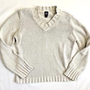 May include: A light beige long-sleeve sweater with a V-neckline and ribbed cuffs and hem. The knit garment features a visible Gap brand label in black and white at the inner collar. The texture is a consistent, medium-gauge knit pattern.