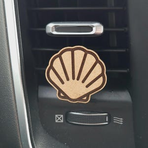 May include: A wooden car air freshener shaped like a seashell. The seashell is light brown and has a detailed design. It is hanging from a car's air vent.