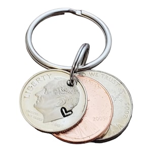 May include: A silver keychain with three coins attached. The coins are a silver dime with a heart cutout, a copper penny, and a silver nickel. The dime and nickel have the words "Liberty" and "In God We Trust" on them. The penny has the words "We Trust" and "Liberty" on it. The coins are dated 2005 and 2002.