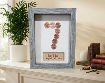 May include: A rectangular shadow box frame with a weathered gray finish containing a white background. Copper-colored coins are arranged to form the number seven. Below the coins, a rectangular plaque displays the text Tony & Sue August 7th, 2015.