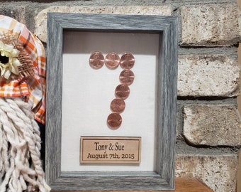 May include: A rectangular shadow box frame with a gray wood-grain finish containing a white background. Seven copper-colored pennies are arranged to form the number seven. Below the pennies is a rectangular wooden plaque with the text Tony & Sue August 7th, 2015.