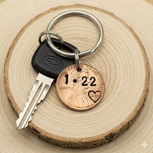 May include: A copper penny keychain with the date "1.22" and a heart cutout. The penny is attached to a silver key ring with a car key. The keychain is displayed on a wooden slice.