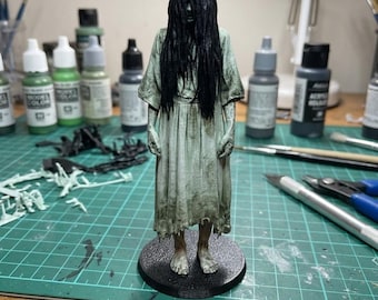 May include: A miniature figurine of a figure with long, straight black hair covering the face, wearing a tattered, pale green dress. The figure stands barefoot on a circular black base, featuring a weathered, textured appearance with visible skin details.