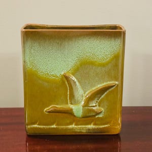 May include: A square ceramic vase with a flying bird relief. The vase has a two-tone glaze, with a light green top and a golden-brown base. The bird is a darker brown colour. The vase is sitting on a dark brown surface.