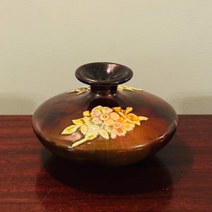 May include: A brown ceramic vase with a dark brown top and a floral design. The flowers are in shades of yellow, orange, and pink, with green leaves. The vase is on a dark wood surface.