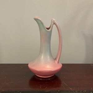 Puede incluir: Una jarra de cerámica esmaltada rosa y azul con un asa curva. La jarra tiene una base ancha y un cuello estrecho.