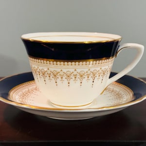 Puede incluir: Una taza de té y un platillo blancos con un borde dorado y un patrón floral azul y dorado. La taza y el platillo están sobre una superficie marrón oscura.