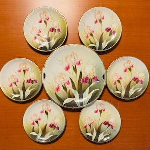 Può includere: Un set di sette piatti rotondi in porcellana con un motivo floreale di iris dipinto a mano in tonalità crema, rosa e viola intenso su uno sfondo verde tenue. Ogni piatto è rifinito con un delicato bordo dorato lungo il profilo circolare.