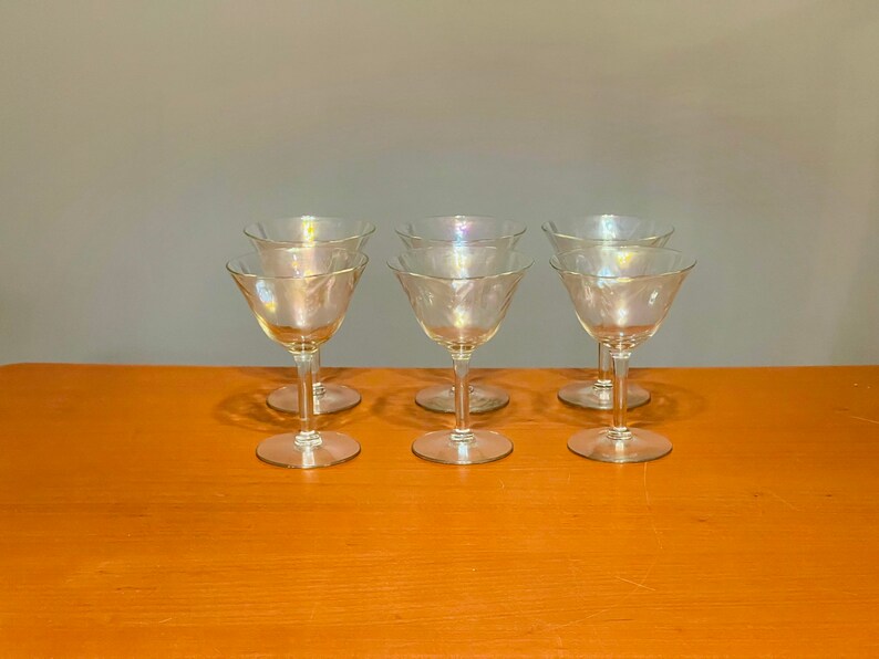 May include: Six clear glass stemmed drinking glasses with a delicate etched design. The glasses are arranged in two rows of three on a wooden surface.