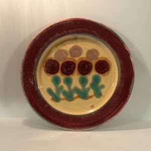 May include: A round ceramic plate with a wide, textured dark red rim. The center features a cream-colored base decorated with a hand-painted design of four teal green stems, four dark red circular flower heads, and three light brown circles.