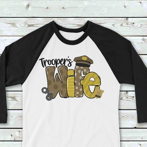 May include: A white and black baseball tee shirt with a brown and yellow graphic that says "Trooper's Wife". The graphic includes a police officer's hat, handcuffs, and a badge.