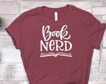 May include: A maroon t-shirt with the words "Book Nerd" in white script and a book graphic. The shirt is styled with sunglasses and small potted plants on a light wood-grain surface.
