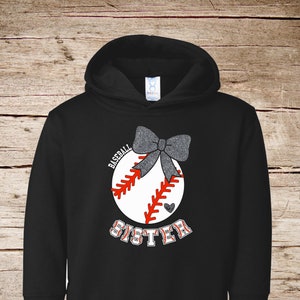 Op de afbeelding: Zwart hoodie met een witte baseball afbeelding en de tekst "Baseball Sister" in wit met een grijze glitter strik bovenop de baseball.
