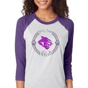 May include: White and purple raglan baseball tee with a Wildcats logo. The logo features a purple glittery wildcat head inside a circle with paw prints and the words "your team" and "wildcats".