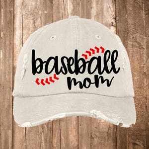 K&ouml;nnte beinhalten: Eine wei&szlig;e Baseballkappe mit ausgefransten Kanten und dem Text "baseball mom" in Schwarz mit roten N&auml;hten.
