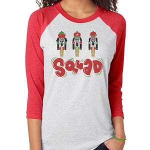 May include: A white and red raglan sleeve shirt featuring three cartoon nutcrackers and the word "Squad" in red and green. The nutcrackers are black, green, and red, with various hat designs.