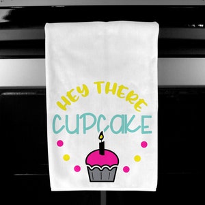 May include: White kitchen towel with the text "HEY THERE CUPCAKE" in yellow and teal. A pink cupcake with a lit candle is below the text, surrounded by pink and yellow dots. The towel is hanging on a black oven.