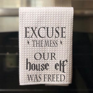 May include: White kitchen towel with the text "Excuse the mess our house elf was freed" printed in black.
