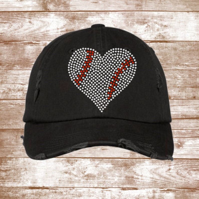 Op de afbeelding: Een zwarte, verweerde baseballpet met een wit hartvormig ontwerp met rode honkbalnaden. Het hart is bedekt met strass.