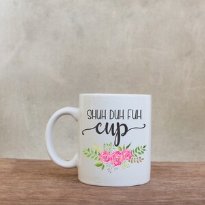 May include: White ceramic mug with the text "SHUH DUH FUH CUP" in black script. The mug features a floral design with pink roses and green leaves. The mug has a white handle and is set on a wooden surface.