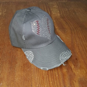 Op de afbeelding: Een grijze baseballpet met een distressed look. De voorkant van de pet heeft een hart van strass met een rood baseballsteekontwerp.