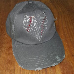 Op de afbeelding: Een grijze, verweerde baseballpet met een strass-baseballhartontwerp. Het hart is rood omrand.