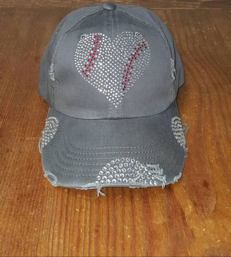 Op de afbeelding: Een grijze baseballpet met een verweerde look. De pet heeft een strass-hartontwerp met rode stiksels. Het hart is gecentreerd op de voorkant van de pet.