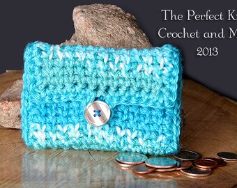 Può includere: Un piccolo porta monete all'uncinetto in un motivo blu e bianco sfumato con un bottone argentato. Il porta monete è su una superficie di legno con diverse monete sparse intorno.
