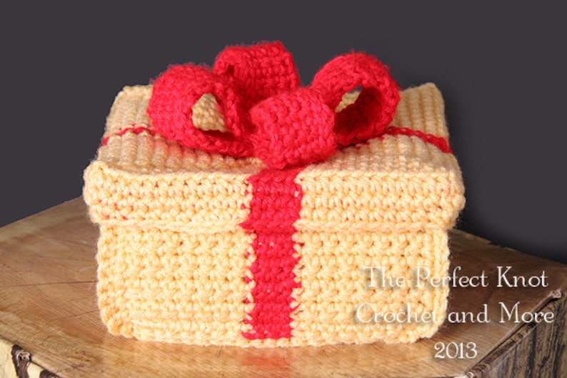 May include: A crocheted gift box with a red bow. The box is made of yellow yarn and has a red stripe down the center. The box is sitting on a wooden surface. The Perfect Knot Crochet and More 2013.
