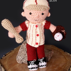 Könnte beinhalten: Eine gehäkelte Baseballspieler-Puppe, die ein rot-weiß gestreiftes Trikot, rote Hosen und einen braunen Baseballhandschuh trägt. Die Puppe hält einen Holzbaseballschläger und steht auf einem Holzstumpf. Die Augen der Puppe sind schwarz und der Mund ist ein kleines Lächeln. Die Puppe trägt eine braune Baseballkappe mit weißem Rand.