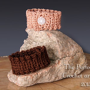 Könnte beinhalten: Zwei gehäkelte Armbänder, eines braun und eines pfirsichfarben, werden auf einem Stein präsentiert. Das pfirsichfarbene Armband hat einen weißen gehäkelten Knopf. Der Text "The Perfect Knot Crochet and More 2013" ist unter dem Stein sichtbar.