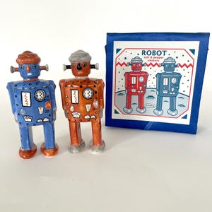 May include: A pair of robot-shaped salt and pepper shakers in blue and orange, with silver accents. The shakers have clock faces and antenna-like features. A blue box with the word "ROBOT" and robot illustrations is also present.