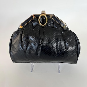 May include: Black clutch with a textured snakeskin pattern. The clasp is gold-toned with a black oval stone in the center. The bag has a rounded shape and is displayed on a clear stand.