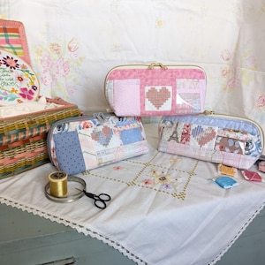 Może przedstawiać: Trzy zapinane na zamek błyskawiczny kosmetyczki z patchworkowej tkaniny z motywem serca. Kosmetyczki są różowe, niebieskie i białe z różnymi wzorami. Kosmetyczki leżą na białym obrusie z haftowanym brzegiem.