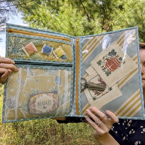 May include: A fabric craft organizer folder featuring a mustard yellow floral print, a white mesh pocket, and a clear vinyl window. The interior includes zippered compartments, three small thread spools, and two embroidered fabric squares.