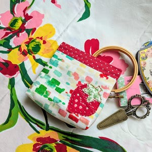 Könnte beinhalten: Eine kleine, rechteckige Stofftasche mit einem roten Erdbeer-Applikation. Die Tasche hat einen weißen Hintergrund mit bunten quadratischen Akzenten und eine rote Tupfenborte. Ein hölzerner Stickrahmen und dekorative Scheren sind ebenfalls sichtbar.