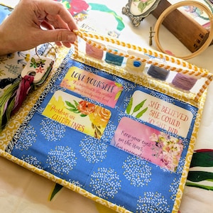 Peut inclure: Un organisateur d'artisanat fait à la main, ouvert, avec un motif floral bleu et une bordure jaune. Il comporte des poches avec des citations inspirantes et contient des bobines de fil coloré. Une main tient le haut de l'organisateur.