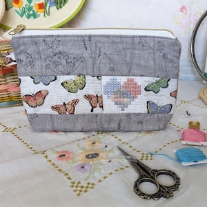 Könnte beinhalten: Eine graue Reißverschlusstasche mit Blumenmuster und Schmetterlingen. Die Tasche hat ein weißes Stoffpanel mit Kreuzstich-Stickerei und Schmetterlingen. Die Tasche liegt auf einer weißen Tischdecke mit gestickten Blumen.