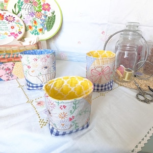 May include: Three small fabric storage baskets with patchwork designs. The baskets are lined with yellow fabric and have a blue and white gingham trim. The baskets are sitting on a white tablecloth with a lace trim.