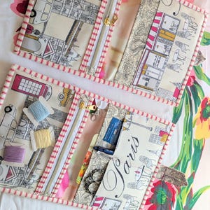 May include: Two open fabric travel sewing kits with zipper closures. The kits feature a cream-colored background with a Parisian street scene print. One kit has spools of thread, the other has scissors and the word "Paris". The edges are trimmed with red and white striped fabric.