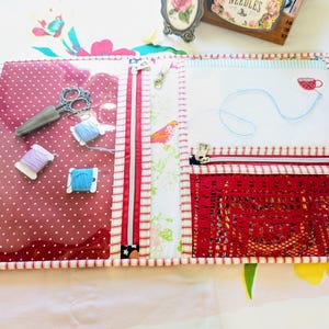 Puede incluir: Un organizador de manualidades transparente con cremallera y secciones de tela de lunares rojos y floral. Contiene tijeras de bordar, carretes de hilo y un diseño de taza de té cosido. El organizador tiene un borde a rayas rojas y blancas.
