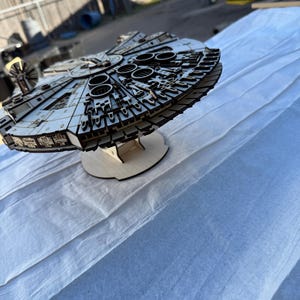 以下が含まれることがあります： ミレニアム・ファルコン宇宙船の詳細な木製モデル。モデルは明るい色で、円形や長方形の要素を含む、複雑なレーザーカットのディテールが施されています。木製のスタンドに置かれ、スターウォーズフランチャイズの象徴的なデザインを披露しています。