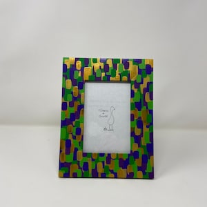 May include: A rectangular picture frame with a colorful, abstract design. The frame is painted with a pattern of green, purple, and gold brushstrokes. The frame has a white interior with the text "Tomato Goose" and a drawing of a goose.