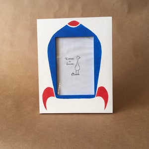 May include: White picture frame with a blue and red design. The frame has a rectangular opening and features the text "Tomato + Goose" with a goose illustration. The frame is on a brown surface.